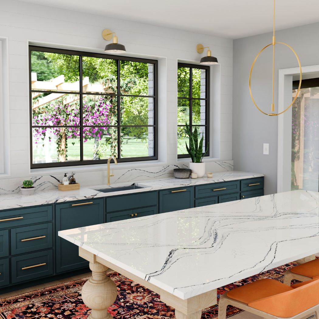 The image is of a modern, spacious kitchen that radiates luxury and elegance. It features white marble countertops, which cover both the central island and the countertops against the wall. The marble texture on these surfaces adds a refined touch. The kitchen cabinetry showcases a teal or blue-green color, providing a cheerful ambiance. A dining area is included within the kitchen space, featuring a dining table accompanied by chairs with bright orange upholstery, which injects a burst of color into the room. There are at least two large windows in the background that allow for ample natural light to illuminate the space, enhancing the airy and open feel of the kitchen. Gold hardware on the cabinets adds a hint of opulence. A vase can be seen among the details of the kitchen, and a potted plant is positioned on one of the windowsills—these elements contribute to the overall aesthetic of the space. A rug is present on the floor, extending from the island, which complements the luxurious feel of the kitchen. The walls of the kitchen are painted in a white shade, which, together with the natural light, contributes to a bright and inviting environment.
